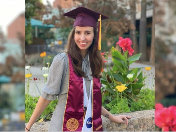 Elinor Saur in graduation cap and sash, posing outside by flowers.